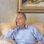 Arnold Brown sitting on a tan couch with a reflective expression.